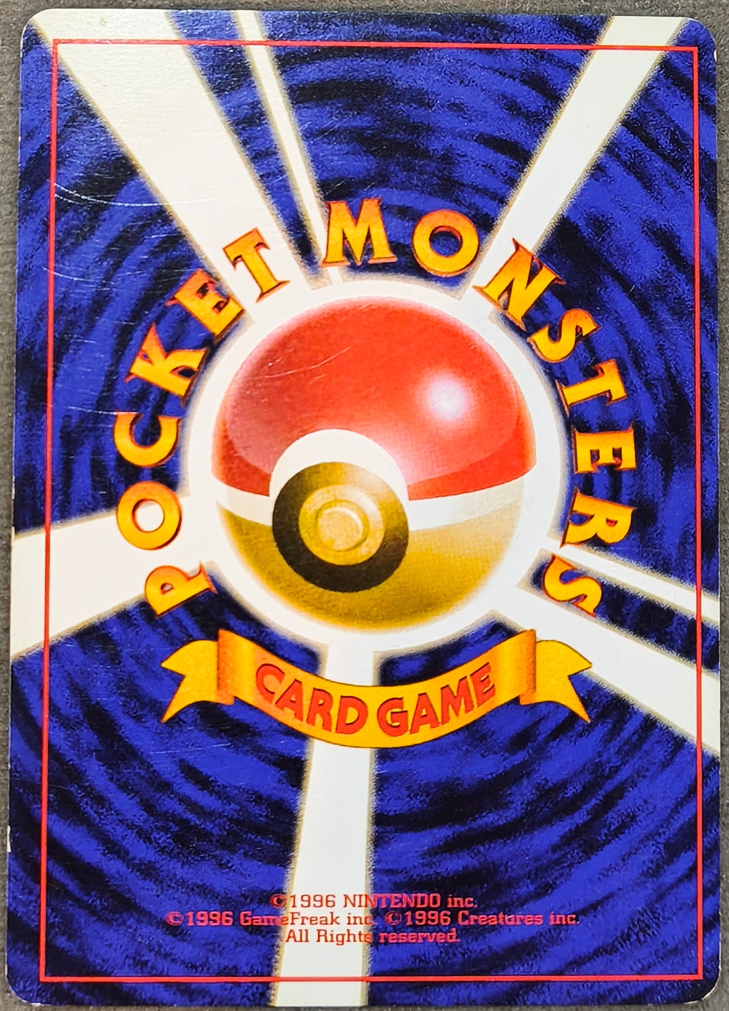 A vintage Pokémon Card Game deck box or card pack photographed from above on a gray fabric surface. The front features the classic blue and white sunburst design with a red-and-yellow Pokémon ball logo in the center and yellow text reading 'Pocket Monsters Card Game.