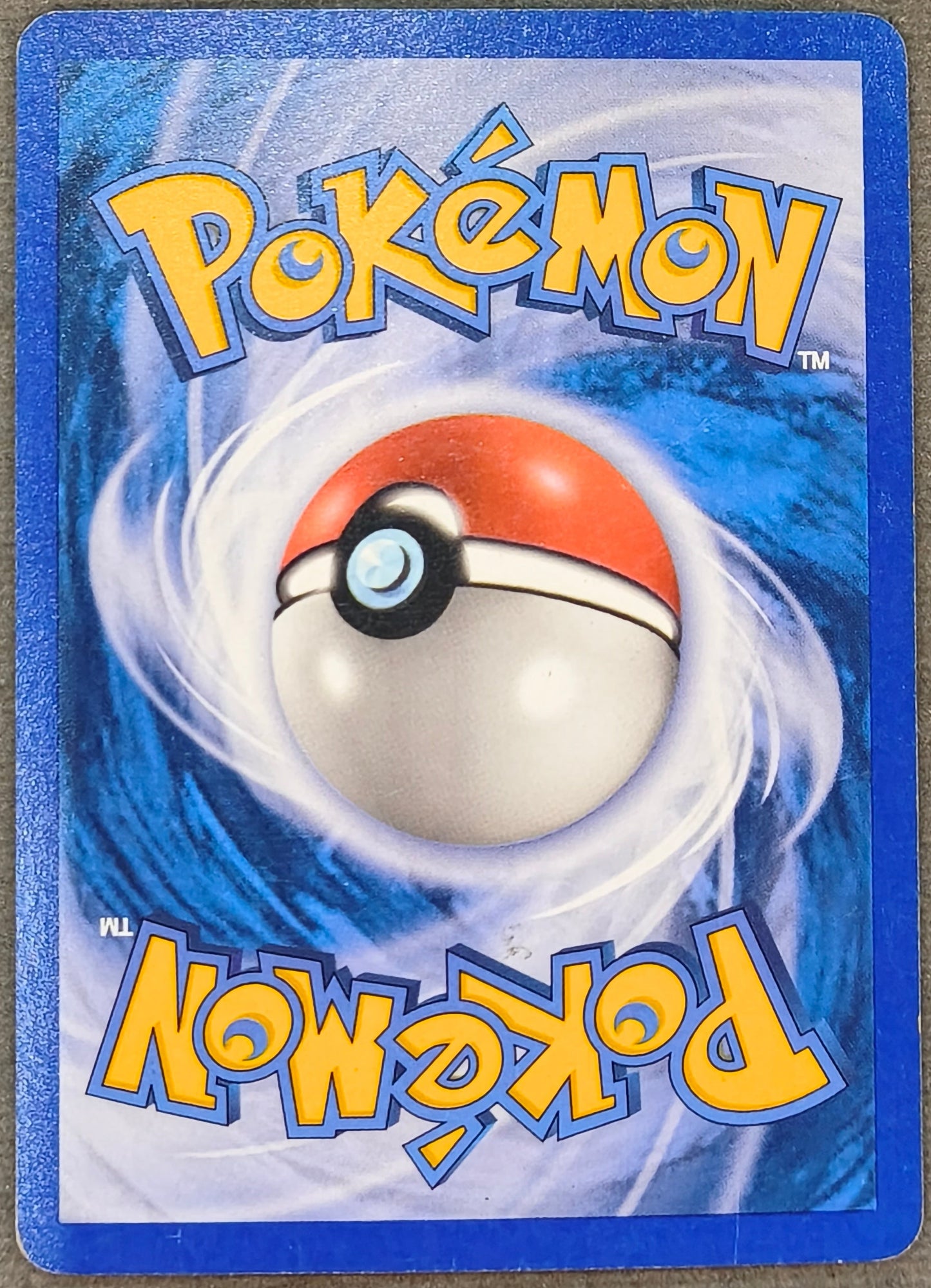 A Pokémon trading card featuring a blue border and a central illustration of a red, white, and black Poké Ball on a swirling blue and white background, with the Pokémon logo in yellow at the top and bottom. The card is photographed on a textured gray surface.